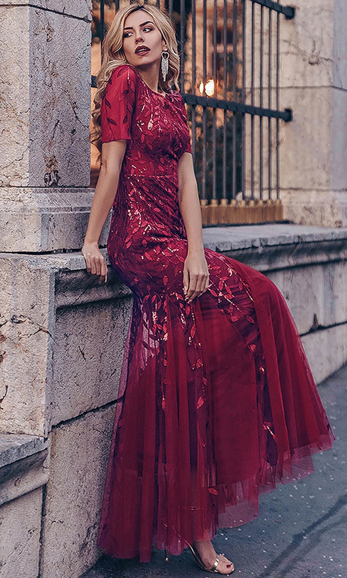 A woman in a Short-Sleeve Modest Long Mermaid Prom Dress 7707 with sheer embroidered floral details sits on a stone ledge in front of a wrought iron fence.